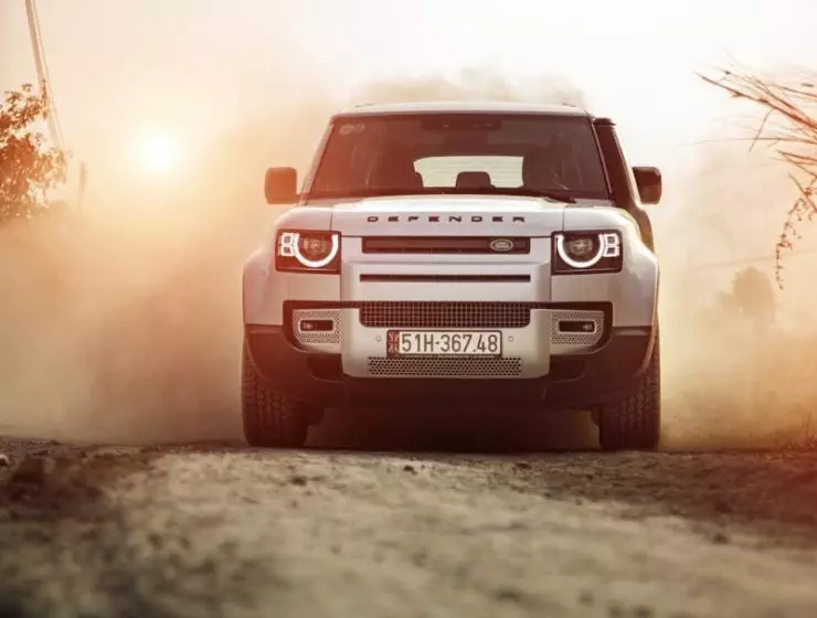 A Land Rover Defender is driving on a dusty road during a sunny sunset.